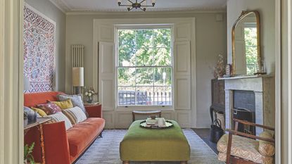 Living room with green ottoman, orange velvet sofa, tapestry on wall and wood shutters, marble fireplace and large mirror above