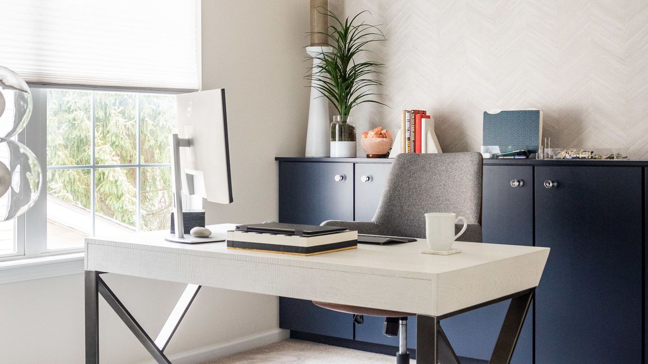 Learning how to make a small office look bigger is useful. Here is a white home office with white walls with a window with white blinds, a blue cabinet with plants and decor on it, a white desk with a computer and white mug on it and black legs