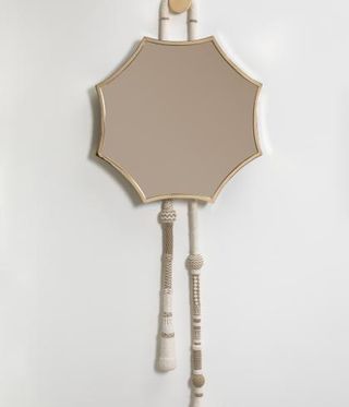 An octagonal mirror hanging on the wall. The mirrored surfaced is made of pink glass, while the mirror also features a golden frame and white cotton tassels at the bottom .