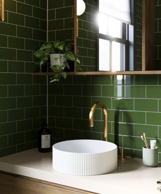 A bathroom sink area with dark green tiles, a mirror with a pothos plant next to it, and a white round basin on a wooden vanity