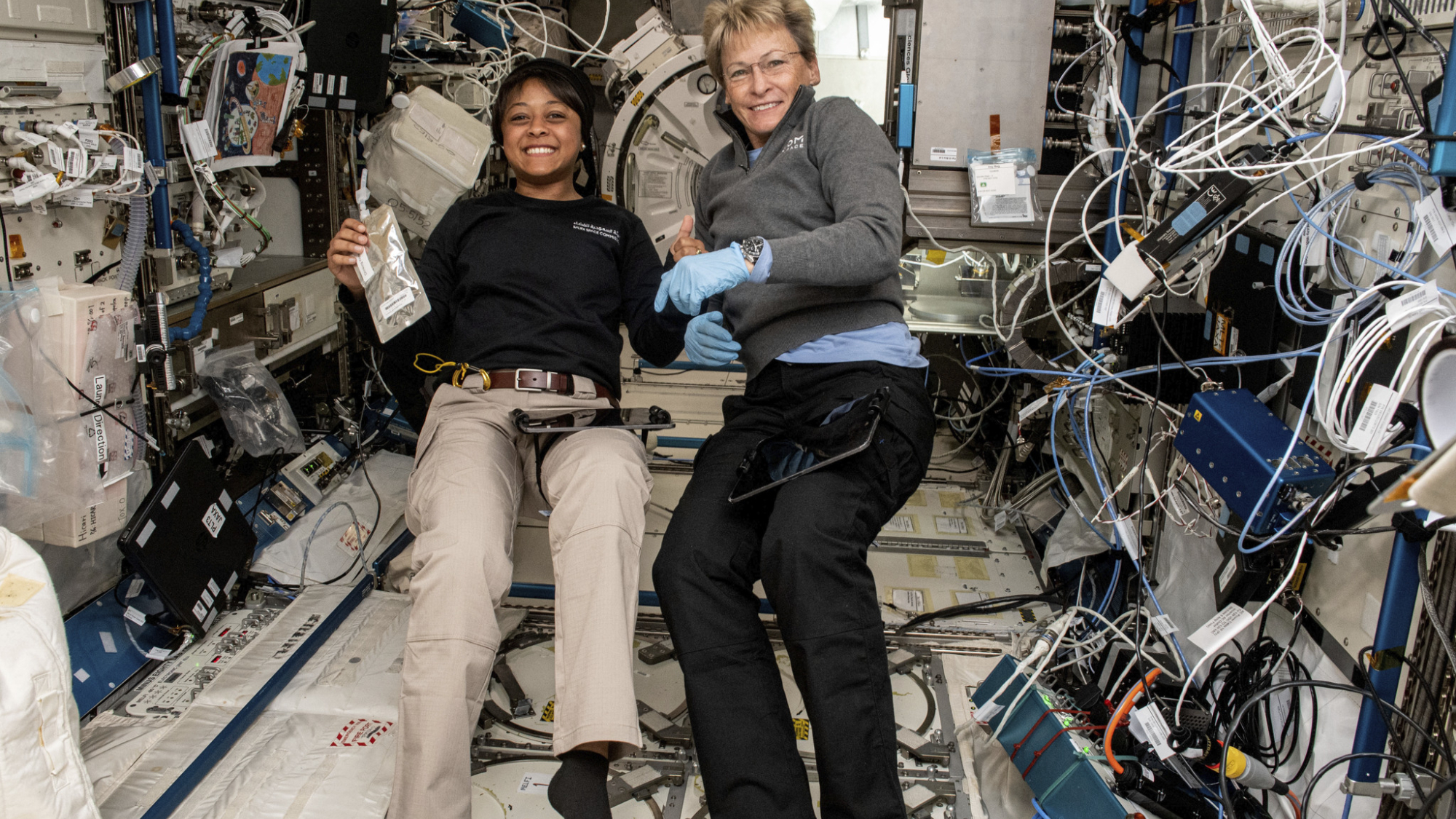 Axiom Space Ax-2 astronauts Rayyanah Barnawi on the left and Ax-2 commander Peggy Whitson smile for a photo surrounded by science gear on International Space Station.