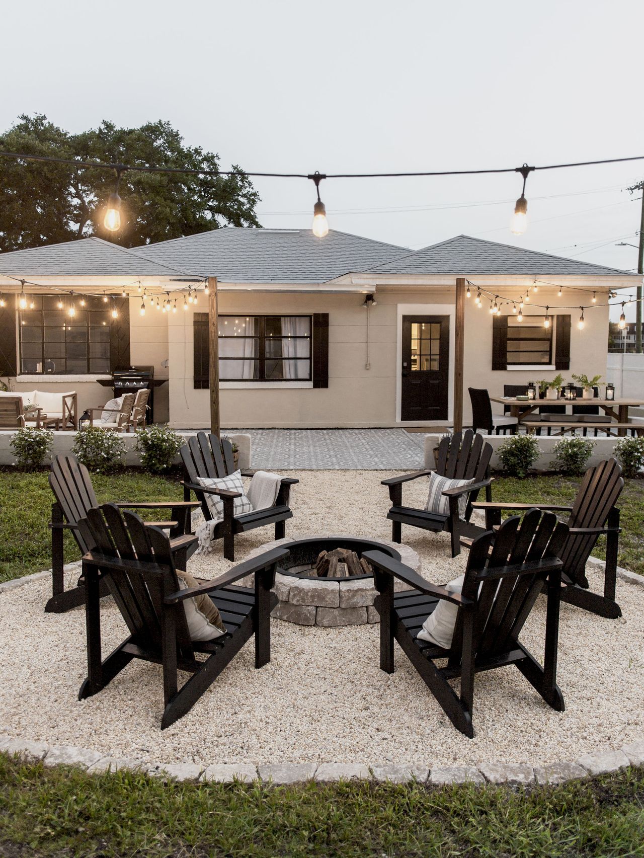 A small seating area outdoors is lit up with string lights with black adirdondack chairs surrounding a firepit in front of the home