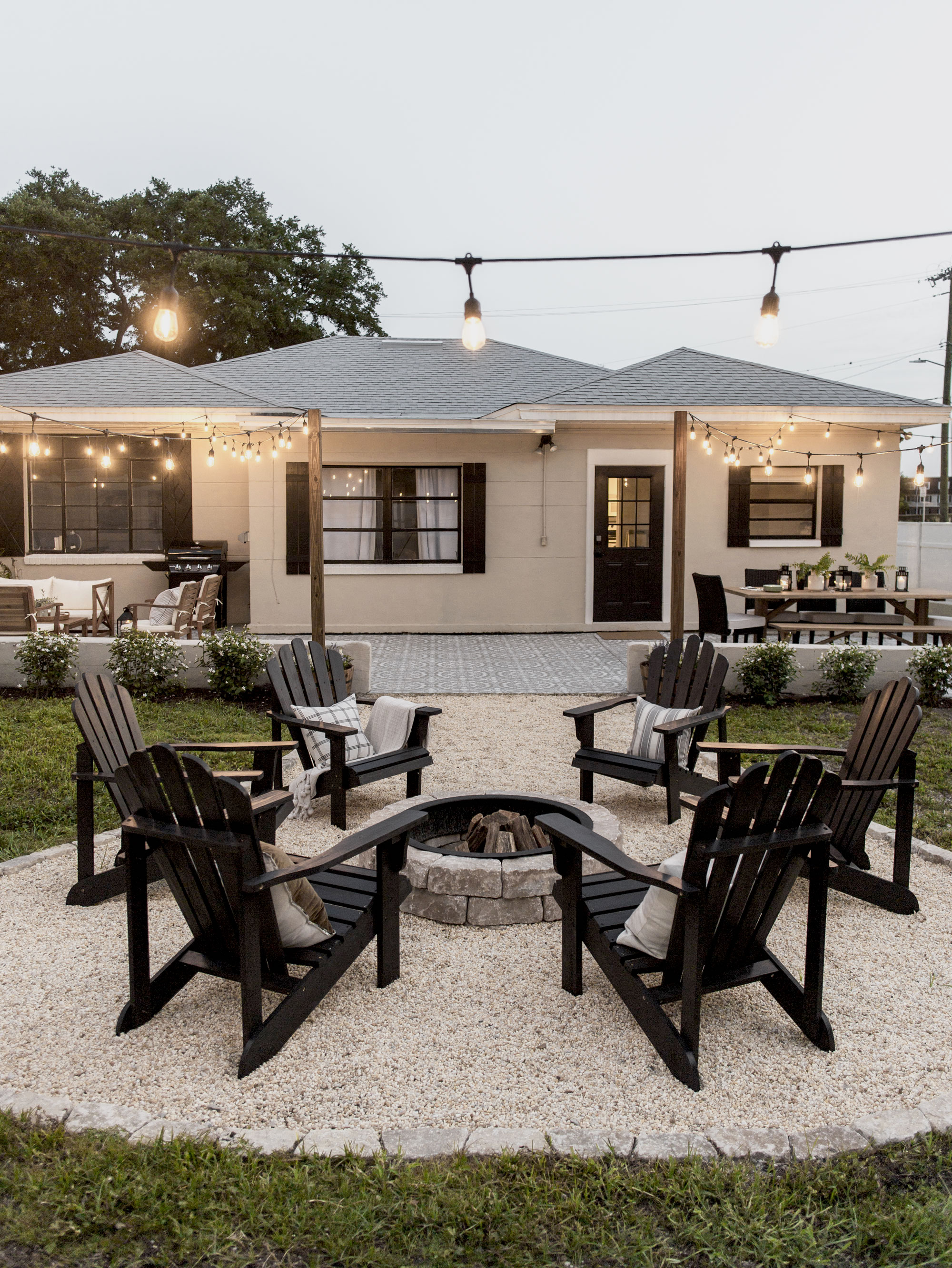A small seating area outdoors is lit up with string lights with black adirdondack chairs surrounding a firepit in front of the home