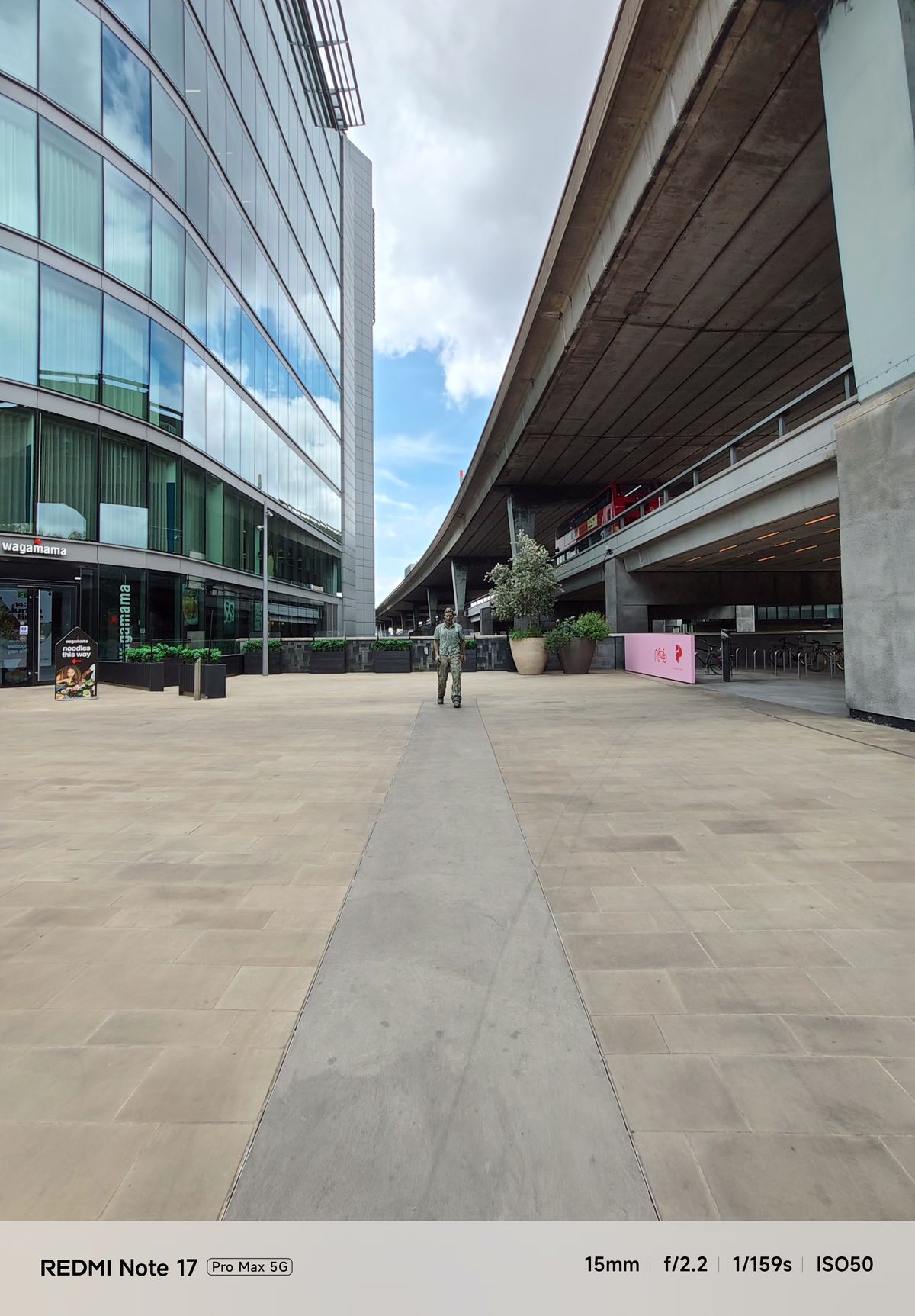 Wide-angle view of an urban plaza with a walking man sculpture beneath an elevated roadway.