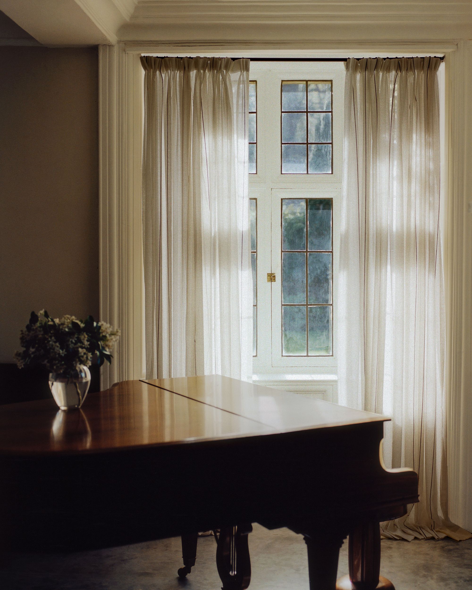 Sheer striped curtains hanging in a window of a dark room. There is a grand piano with a small flower vase on it in the room as well.