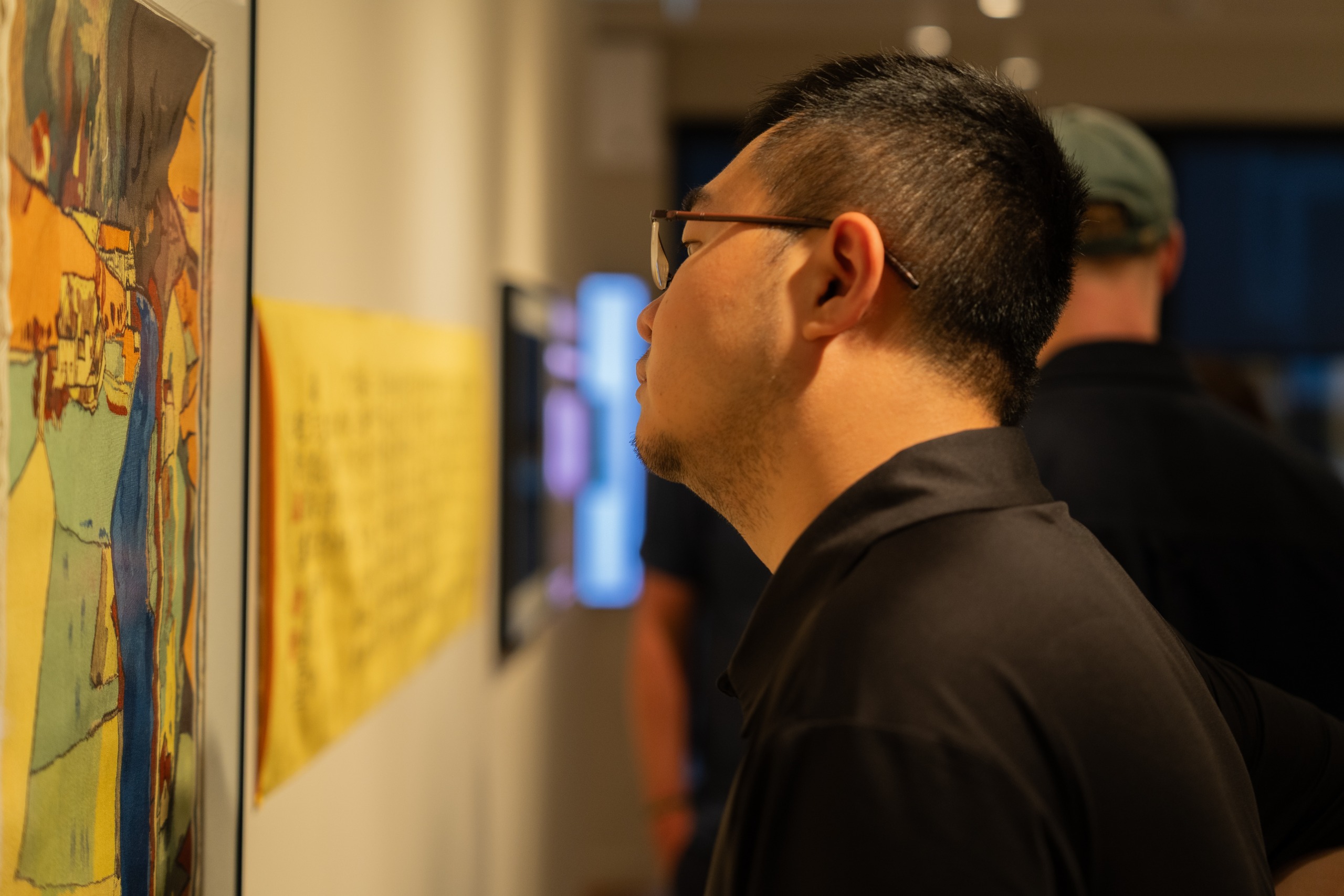 A man in glasses peers closely at a colourful silk tapestry of a landscape in yellows, greens and blues, with a yellow calligraphy panel softly blurred behind it.