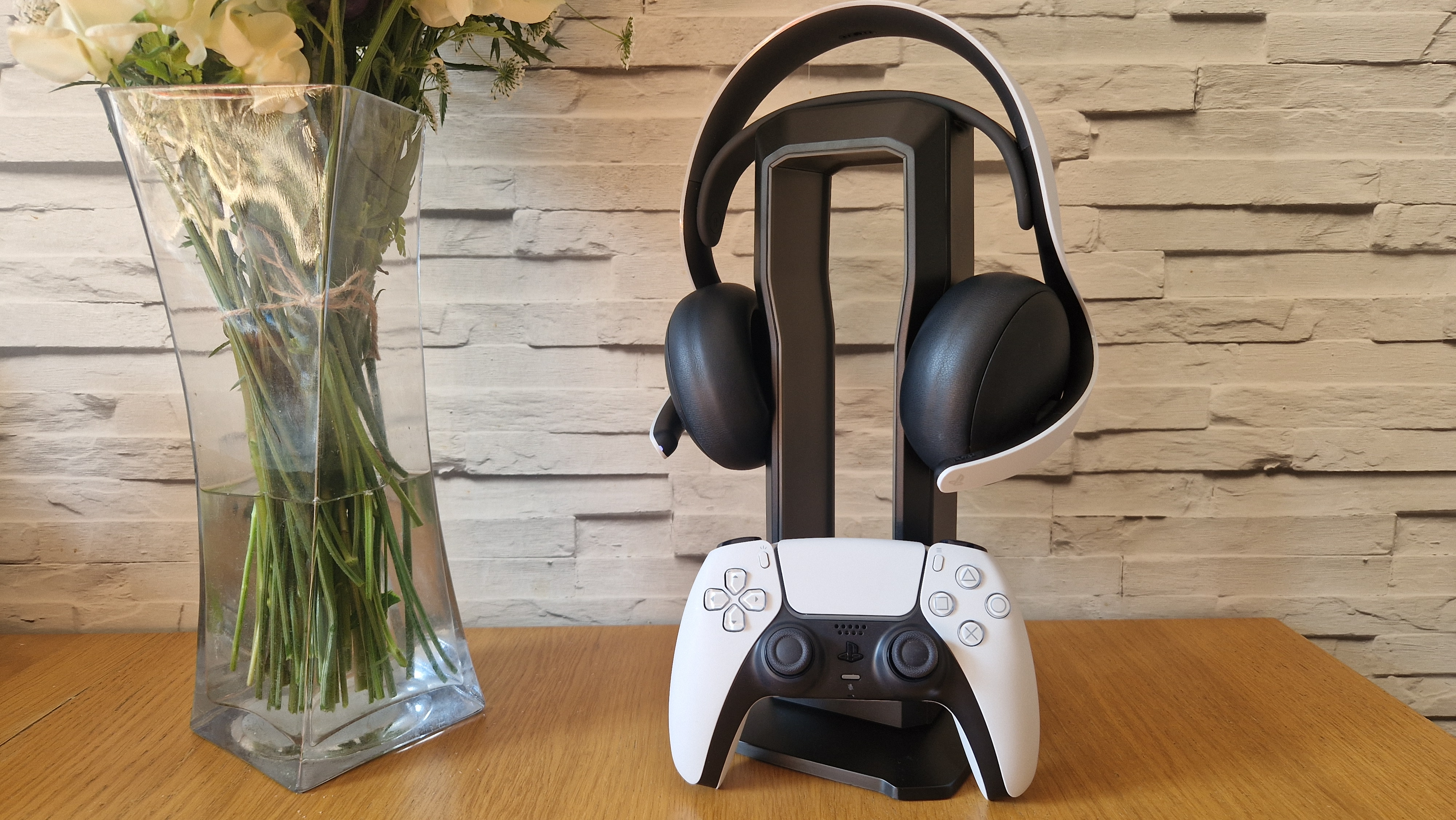 A PlayStation Pulse Elite headset on a headset stand on top of a wooden surface next to a vase and a DualSense controller