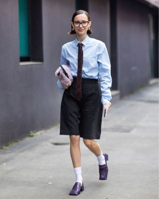 A guest is seen wearing a light blue shirt, grey bermuda shorts, a brown and blue printed tie, white socks, a violet fur bag, gold earrings and purple leather heeled loafers outside Max Mara show during the Milan Fashion Week - Womenswear Fall/Winter 2024-2025 in Milan, Italy.