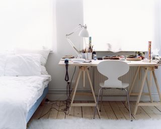 A white small minimalistic bedroom with a white bed and wooden desk next to each other with a fuzzy white rug and modern white chair