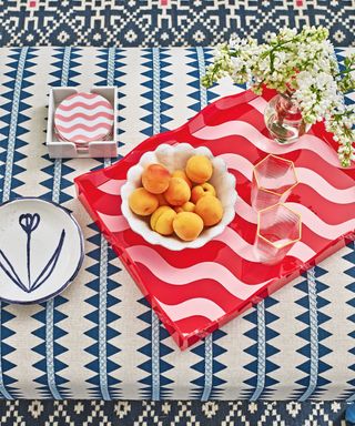 Pink and red striped serving tray, blue patterned tablecloth, plates, coasters, glasses, flowers, bowl of fruit