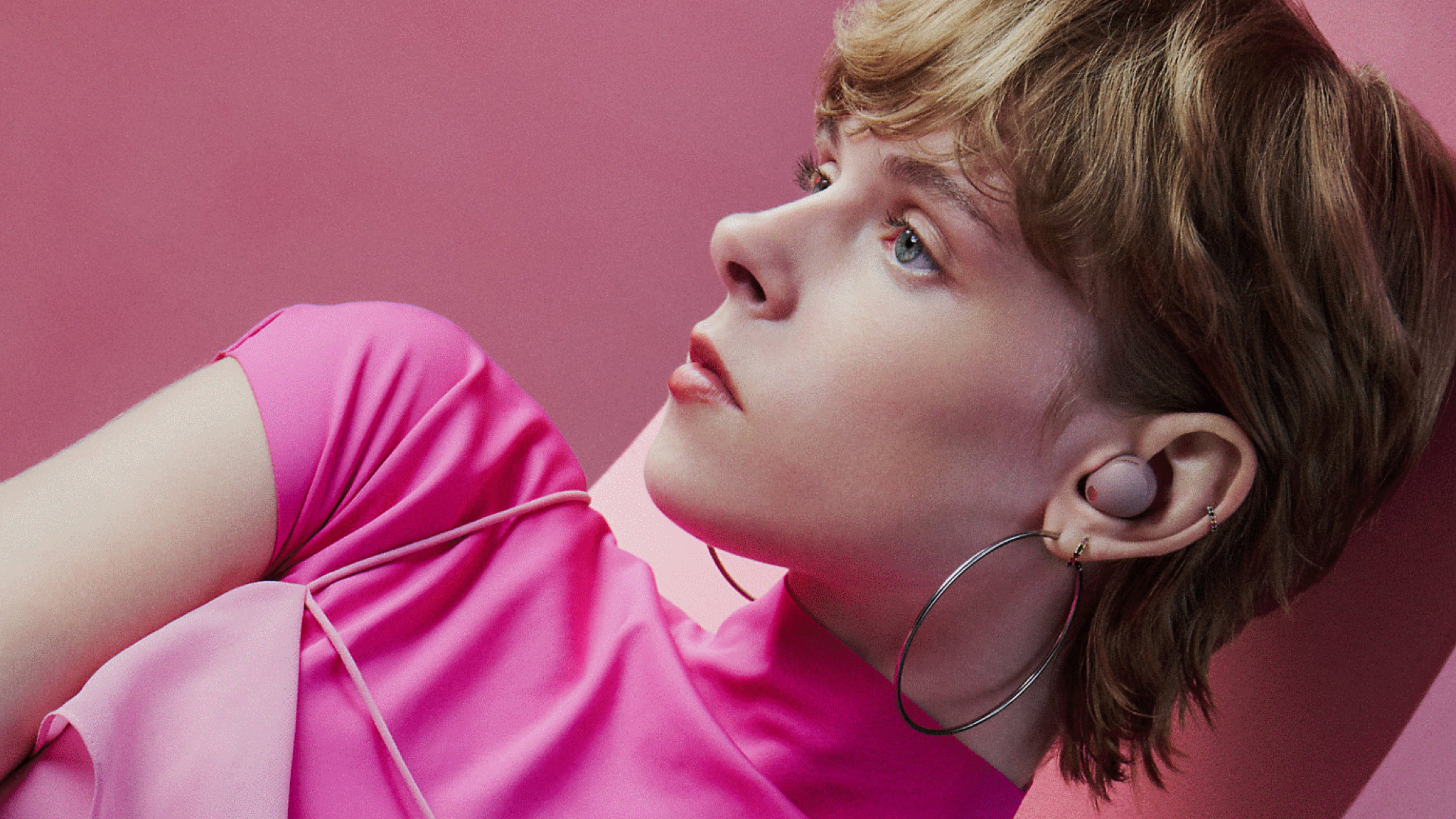 A young woman in pink with blonde hair and large hoop earrings wearing Sony WF-1000XM5 earbud in its pink colour version