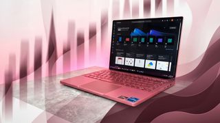 A pink laptop on a gray surface, displaying a dark-themed screen with apps. The background has abstract pink and maroon wave patterns, creating a modern, dynamic feel.