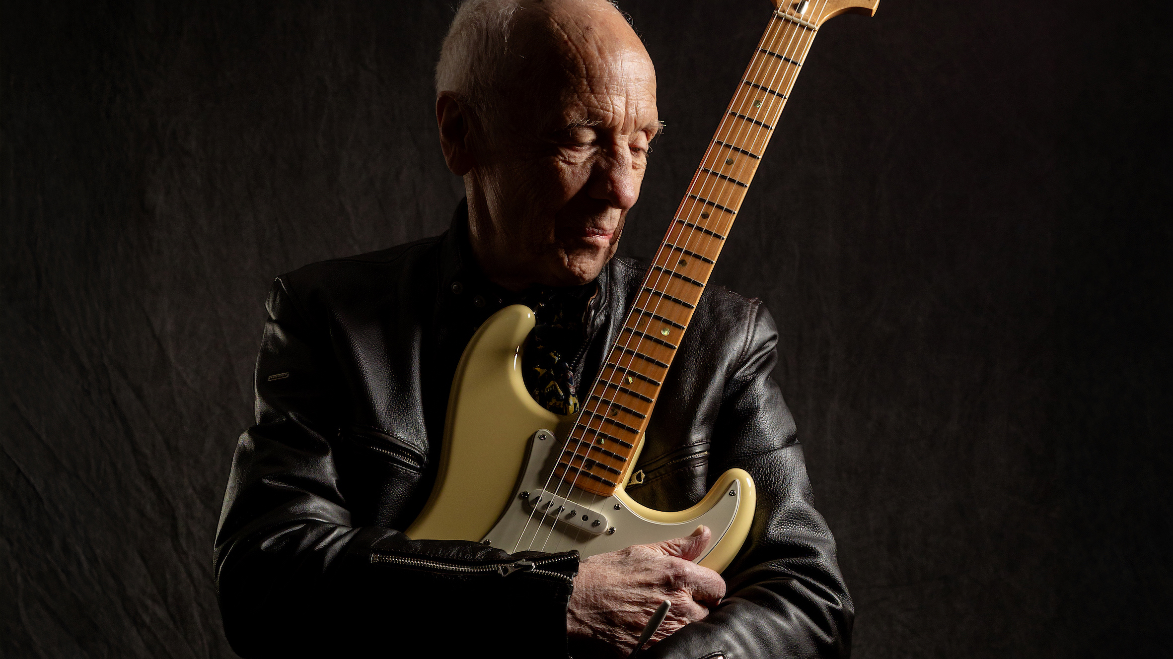 Robin Trower poses in a black leather jacket holding his signature yellow Fender Stratocaster