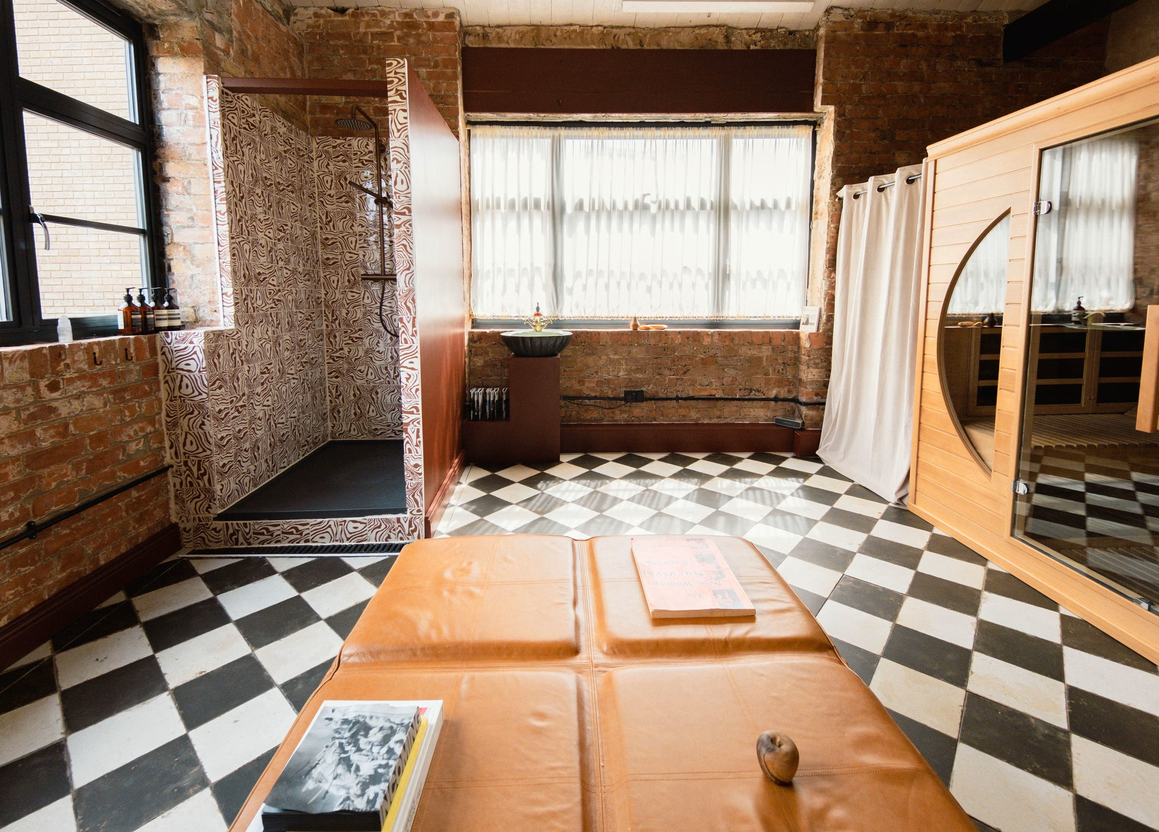 A sauna room with checkered black and white floors, a wooden sauna cabin, raw brick walls, plenty of natural light, and quirky touches like a brown leather banquette and a patterned, rectangular built-in shower.