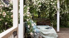 We love spring porch decor ideas. Here is a front porch with a white pergola with white flowers above it and a blue day bed with a light green floral blanket with magazines on it, and a silver watering can