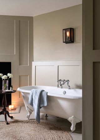 A small light in a glass box above the bath tub. Next to the bath rub is a table and vase with flowers on top.