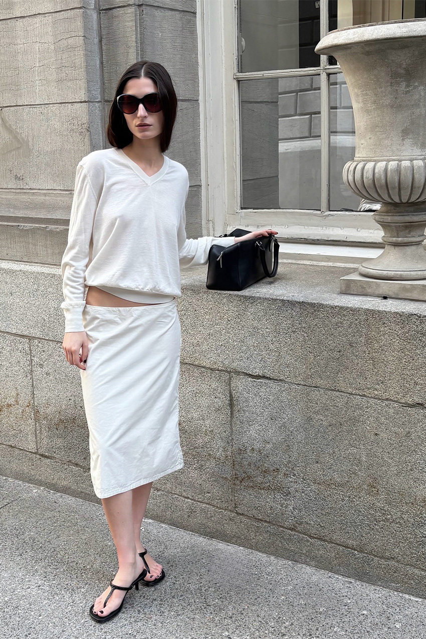 White outfit idea for summer is shown in a photo of a woman standing outside wearing large black sunglasses with a white sweater, a white midi skirt, black thong sandals, a black shoulder bag