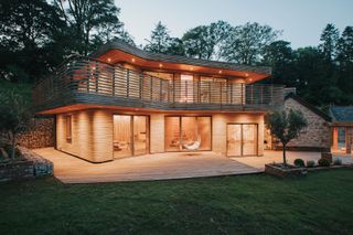 A timber home at night lit up all around the front with a stone house to the side