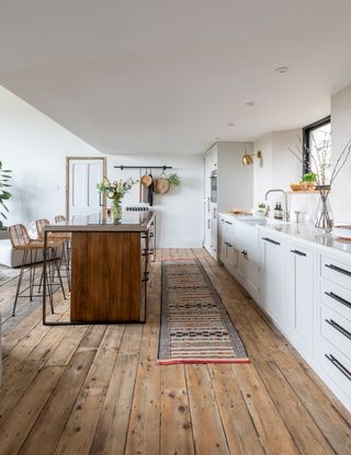 Hardwood plank flooring in open plan modern rustic kitchen with white cabinets, dark wood island and woven bar stools.