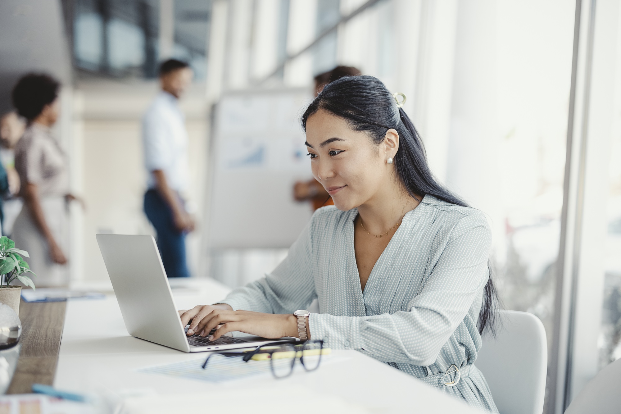 Asian woman working on laptop. Business woman busy working on laptop computer at office with colleagues in the background.