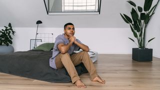 A man sits on a mattress on the floor placed on a hardwood floor so it sleeps cooler