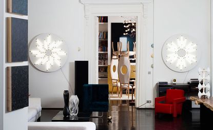 Inside an apartment with a small lounge featuring a white chair, black side table, two circular white lights attached to the wall, a small red chair. The room next door features two tall backed chairs with circular holes in their back centre. 