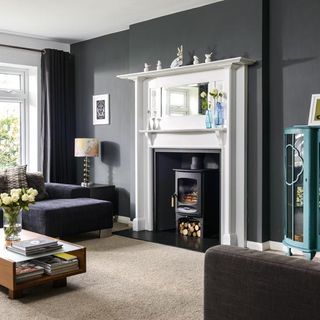 a living room with dark grey walls,, light brown carpet, dark furniture and a woodburner fireplace surrounded by a white mantle