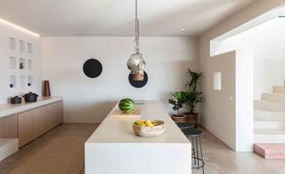 White kitchen with long center and wall counters, round chairs, various cooking equipment and two potted plants.