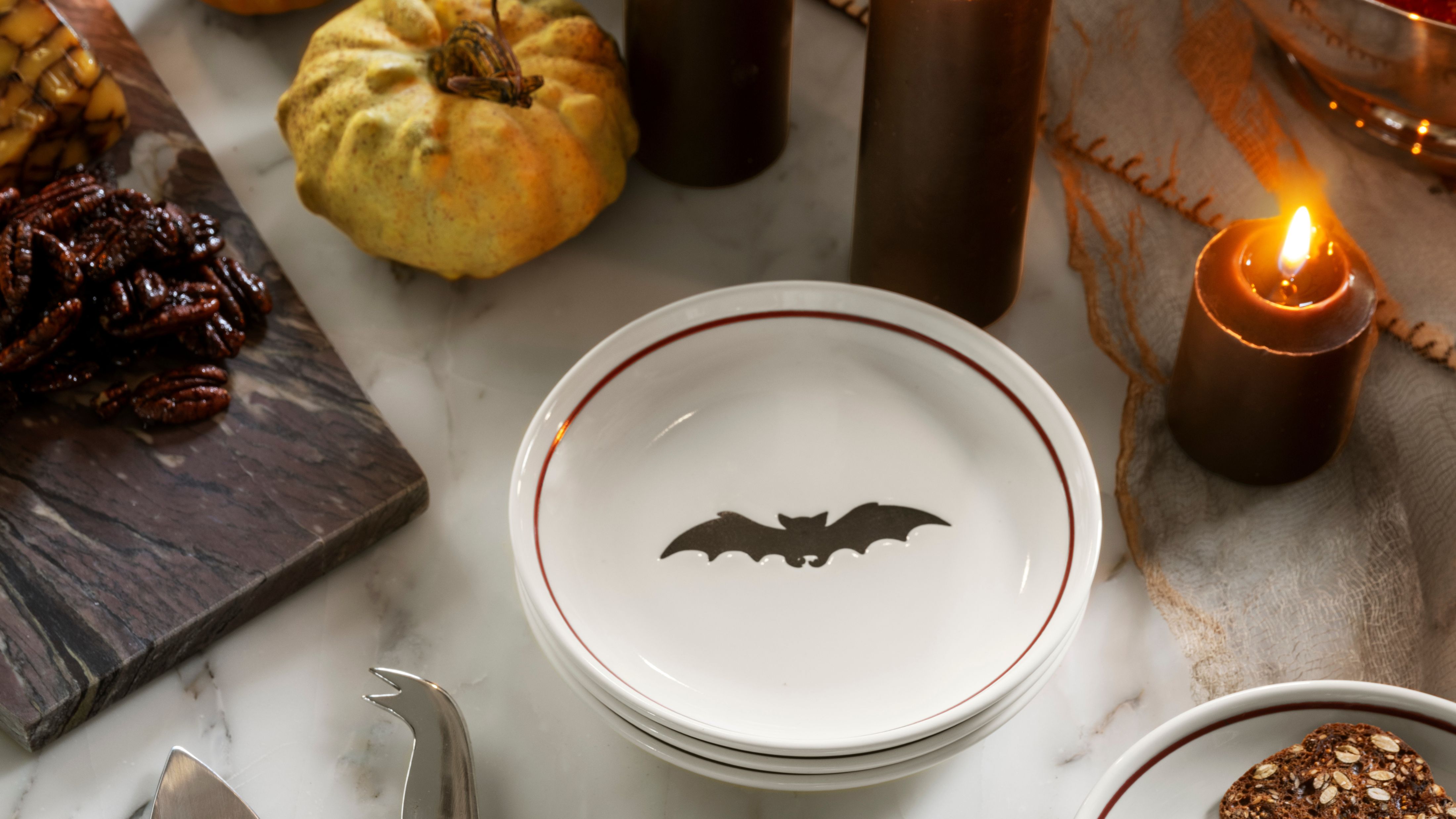 Halloween tablescape featuring bat-motif plates, gourds, and lit taper candles.
