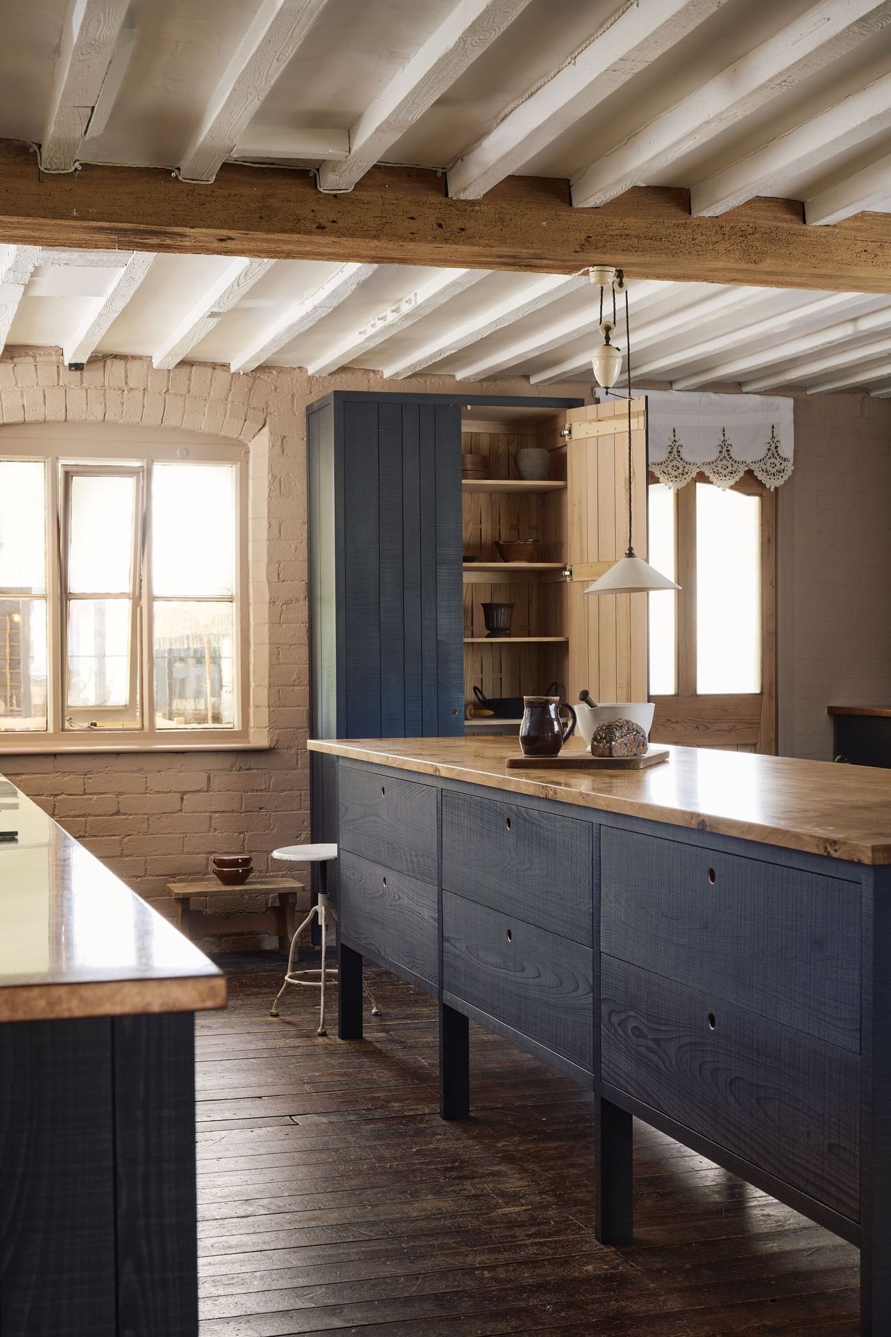 Image of a rustic kitchen countertop, island, and pantry. The floors are a dark wood and the walls are white brick. The cabinets on the island and pantry are stained in a navy blue color and the countertop material is copper. There is a small white pendant light hanging over the island.