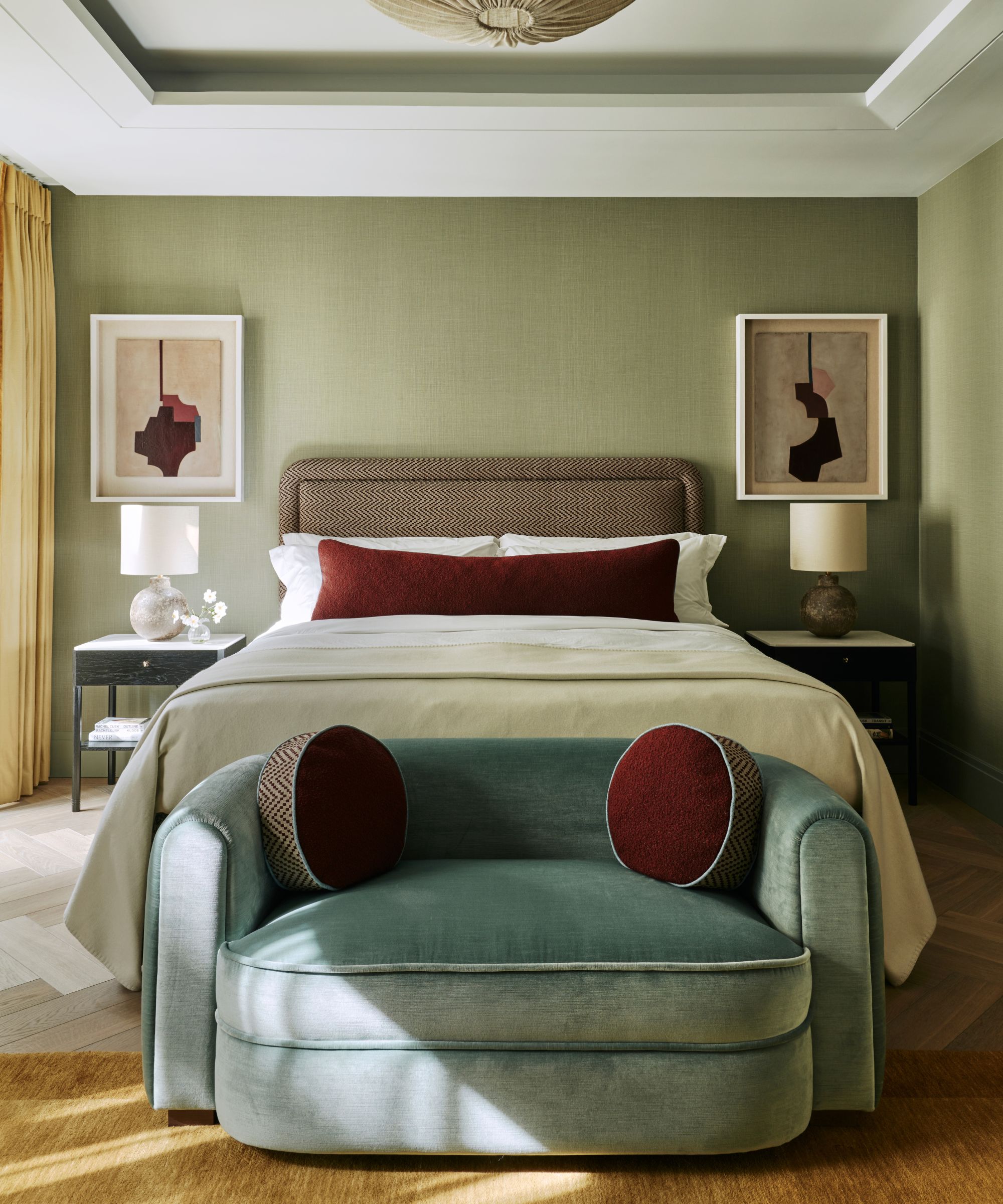 cozy modern earthy green bedroom with green walls, a large neutral bed styled with a red velvet lumbar pillow, a blue velvet small couch at the foot of the bed with round pillows