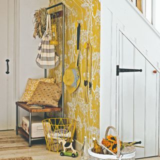 A small hallway with a wall covered in a yellow botanical wallpaper and a storage unit tripling as a coat rack, a bench and shoe storage