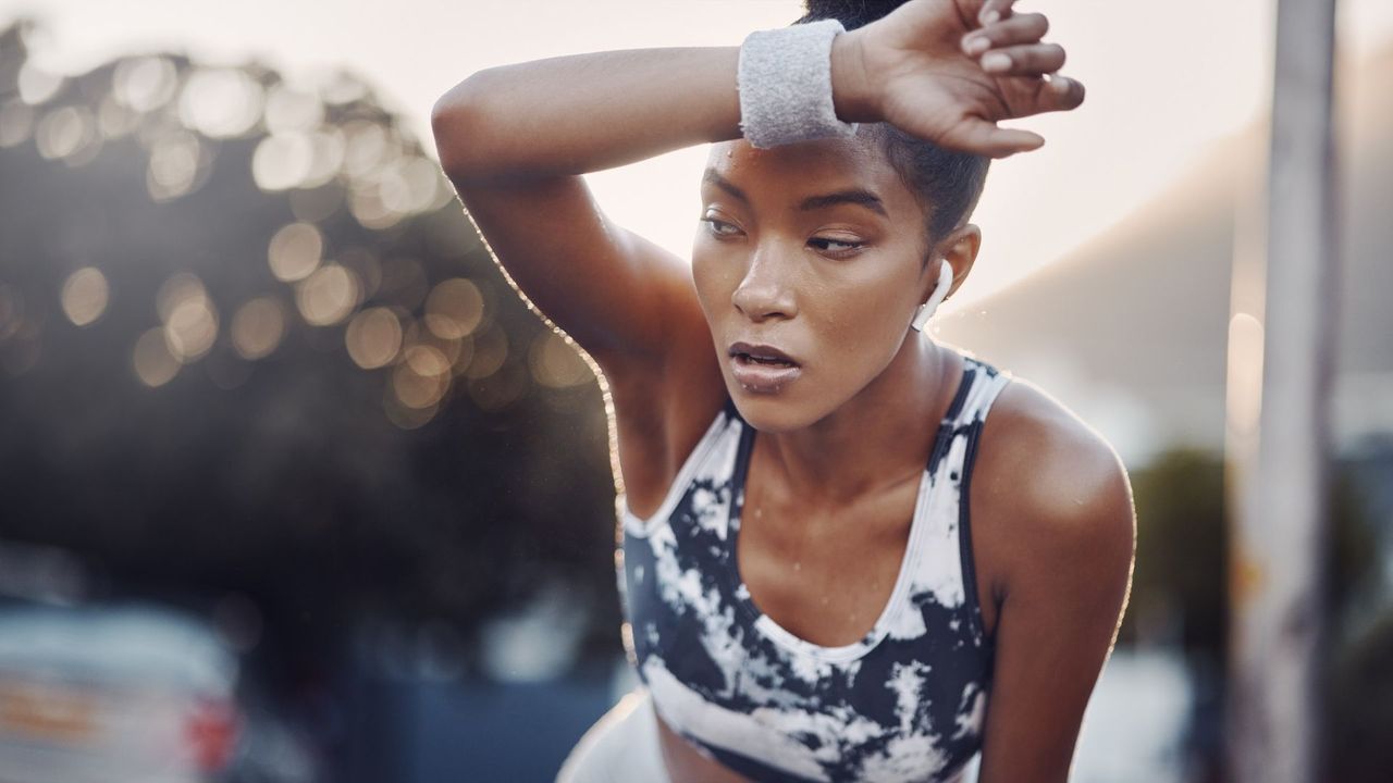 A woman in a tie-dye sports bra wipes her sweating brow with a sweatband. She looks a little unhappy.