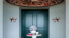 Pale blue entryway with dark teal double doors, two red sconces on walls, a large pink floral ceiling mural, a round, small white table in the centre with a vase and books, and parquet wooden floors
