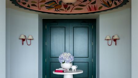 Pale blue entryway with dark teal double doors, two red sconces on walls, a large pink floral ceiling mural, a round, small white table in the centre with a vase and books, and parquet wooden floors