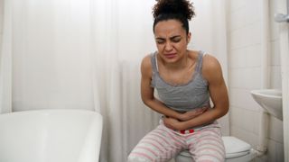 woman with light brown skin sits in a bathroom clutching her stomach as if in pain. She's wearing a tank top and pajama bottoms