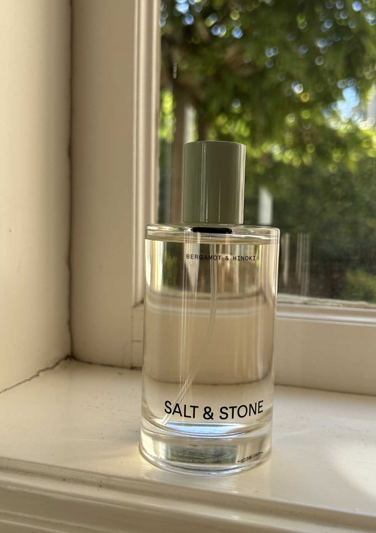 Salt &amp;amp; Stone Bergamot &amp;amp; Hinoki perfume on a sunny windowsill, clear bottle with a muted green cap and trees blurred behind it