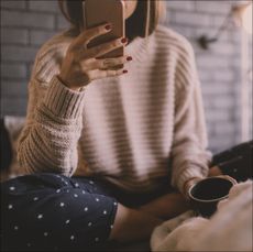 Photo of smiling young woman drinking first morning coffee in her bed, and reading online news on a smart phone