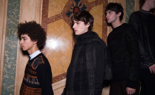 Three male models wearing looks from Pringle of Scotland's collection. One model is wearing a black, orange and white patterned jumper. Another model is wearing a dark coloured piece and black and grey patterned scarf. And the last model is wearing dark brown trousers and a black patterned jumper