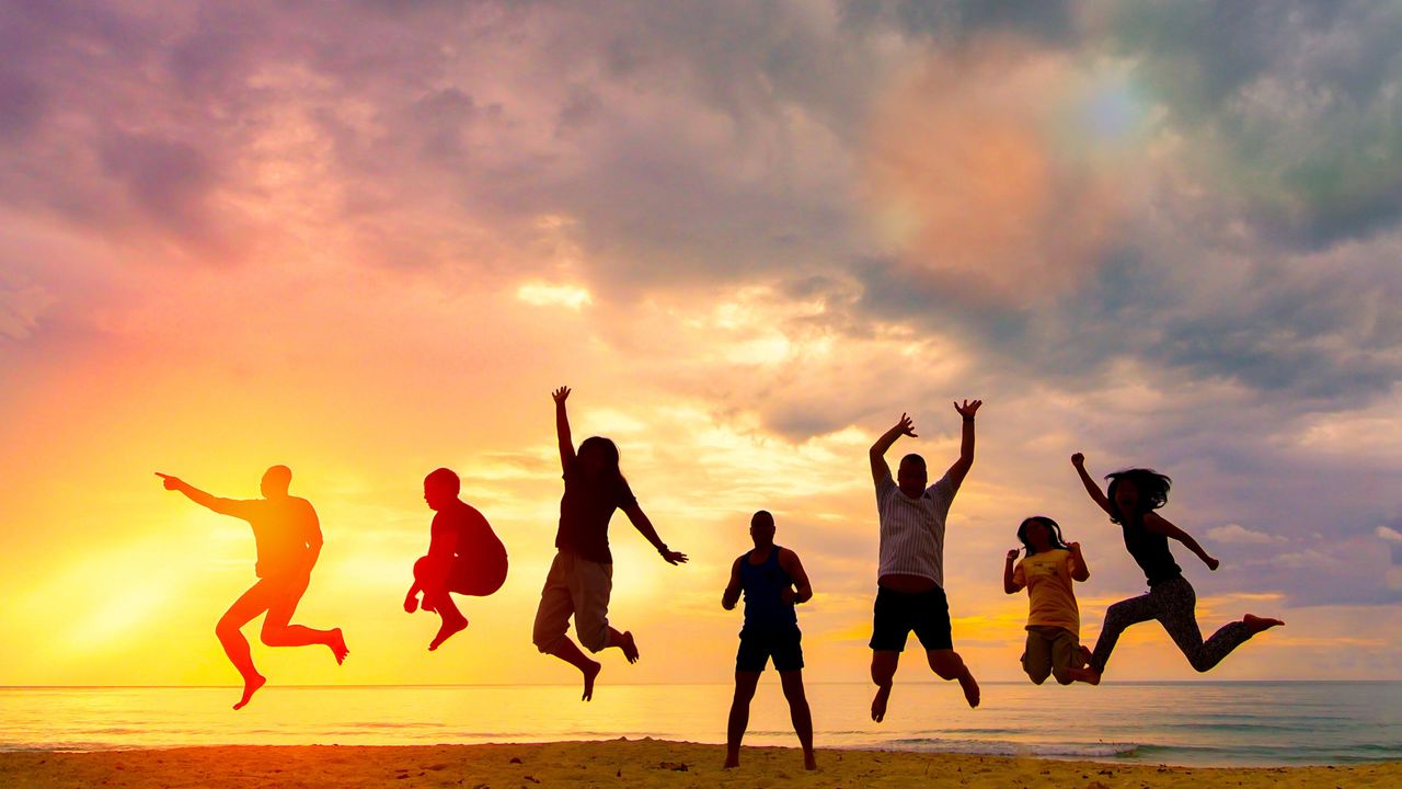 A group of people jump on a beach silhouetted against a sunset.