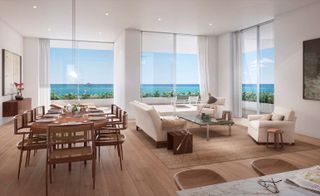 Inside a residence featuring floor-to-ceiling windows overlooking the sea. The interior has a large wooden table with wood chairs to the left side, and beige sofa chairs to the right.