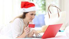 photo of a woman in a santa hat holding a credit card grimaching as she looks at a laptop screen