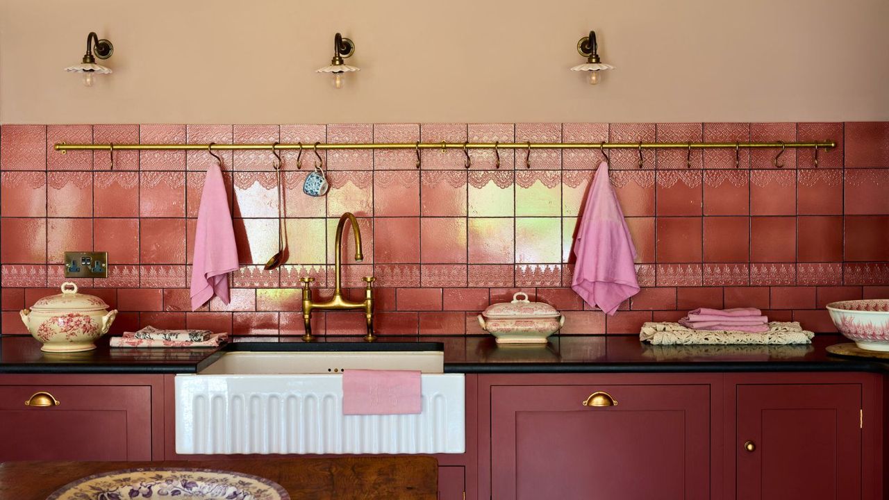 Red kitchen cabinets with brass fixtures, black stone countertops, brass towel rail on pink tiled wall with under mounted white fluted butler sink