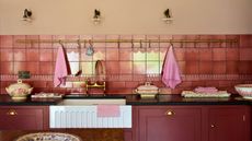 Red kitchen cabinets with brass fixtures, black stone countertops, brass towel rail on pink tiled wall with under mounted white fluted butler sink