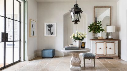 hallway with stone floor tiles, stone circular table with flowers and books, stool, vintage lantern, dresser with mirror, flowers, artwork, crittall doors to left, 