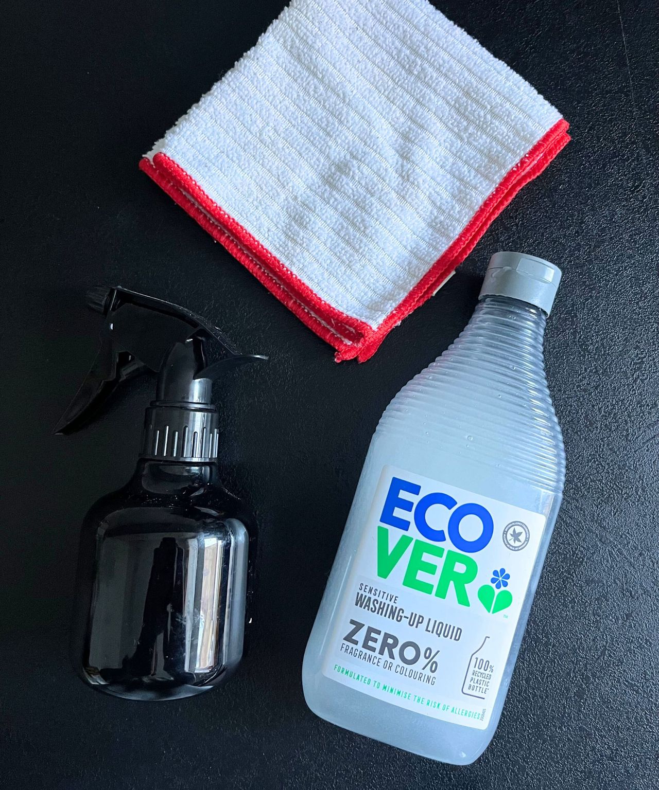 A small black spray bottle filled with vinegar beside a bottle of Ecover washing up liquid. A folded white microfiber cloth above.