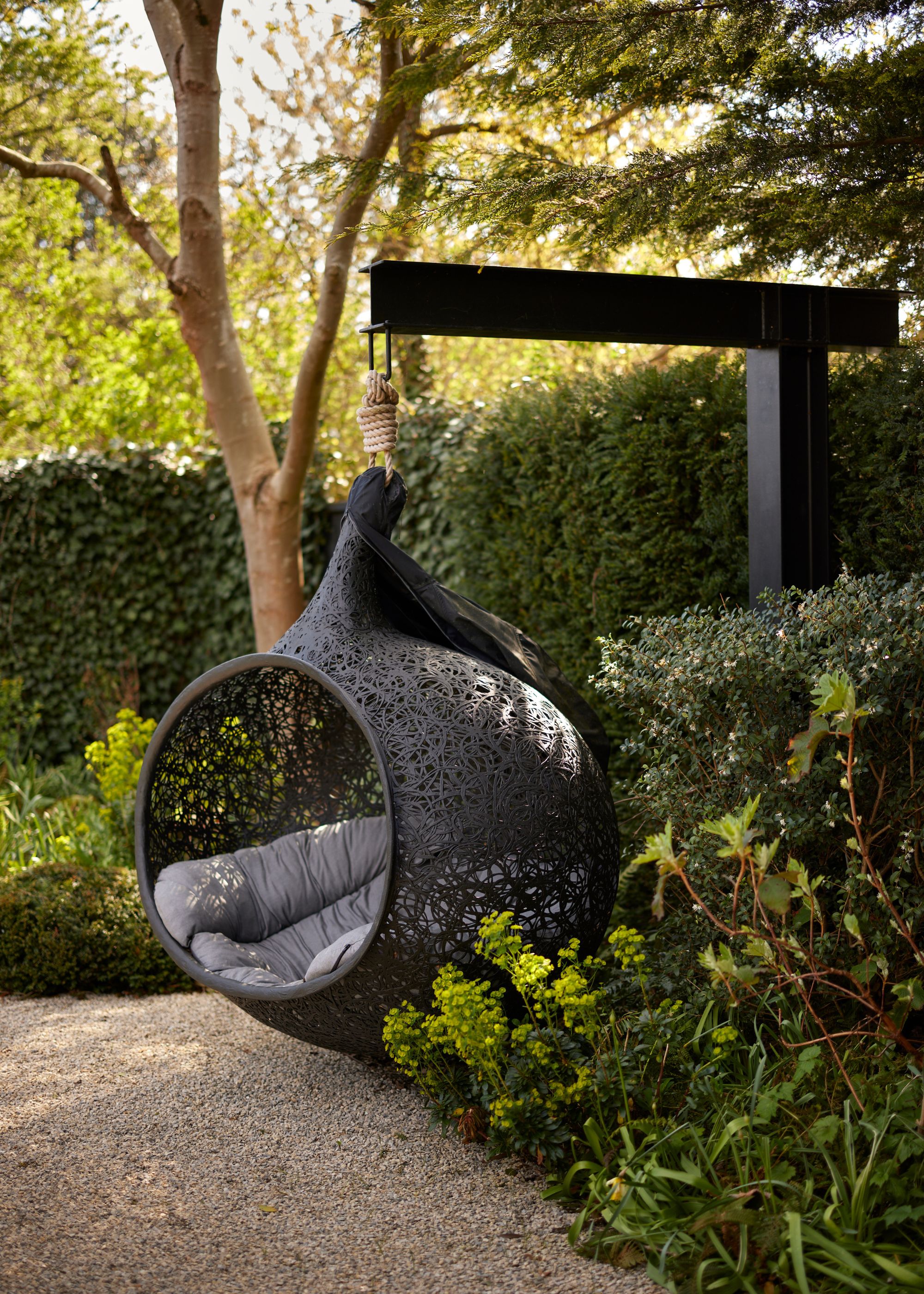 A modern garden with a gravelled ground and lush green planting to the perimeter. A black hanging egg chair positioned within the greenery, suspended from a black frame.