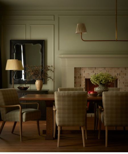 Cozy dining room with dark wooden rectangular dining table, upholstered dining chairs, vase of flowers on the table. Brick fireplace, and sideboard with lamp and vase in background.