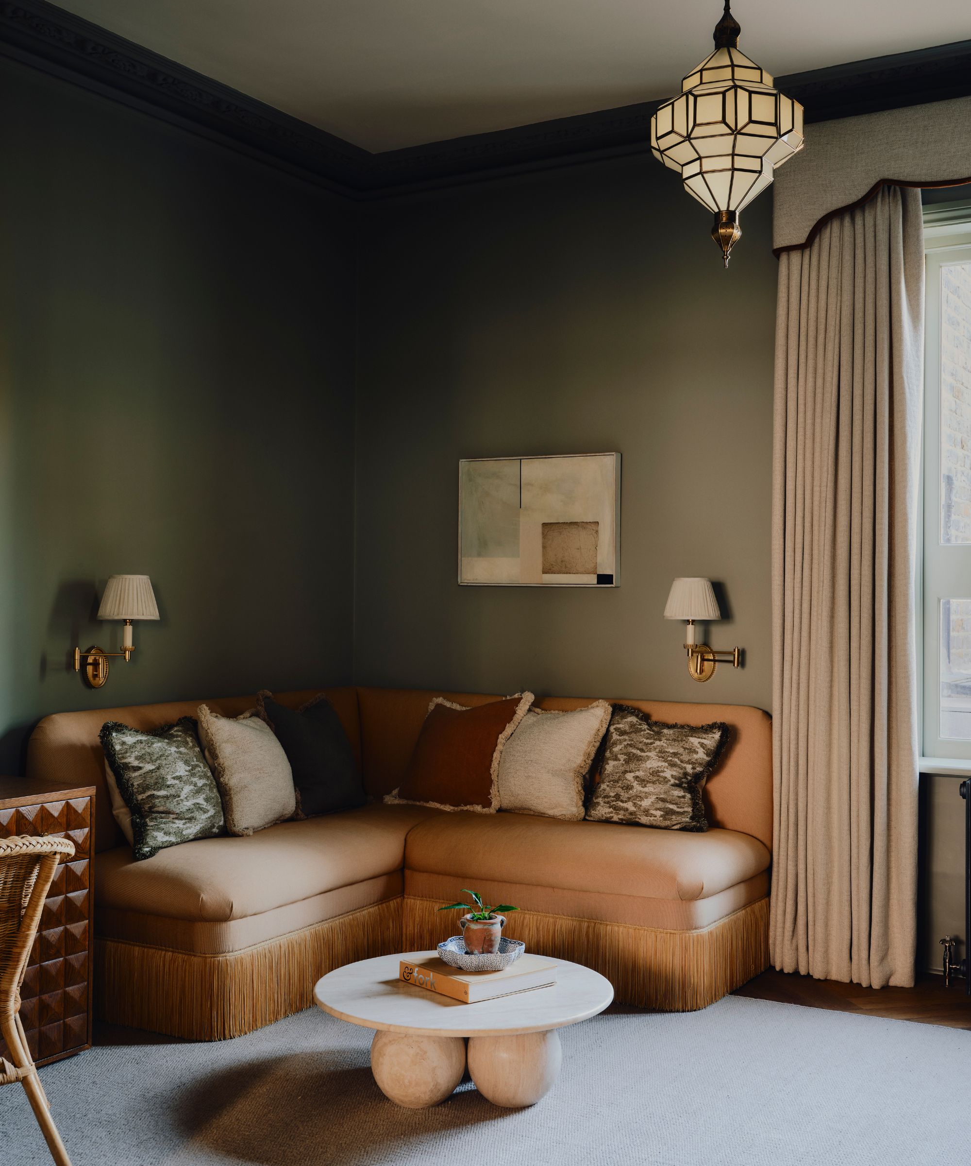 Living room with khaki green walls, pelmet curtains, a brown banquette sofa with fringing, a marble round table, two gold sconces, colourful pillows on top, and a Moroccan lantern light.