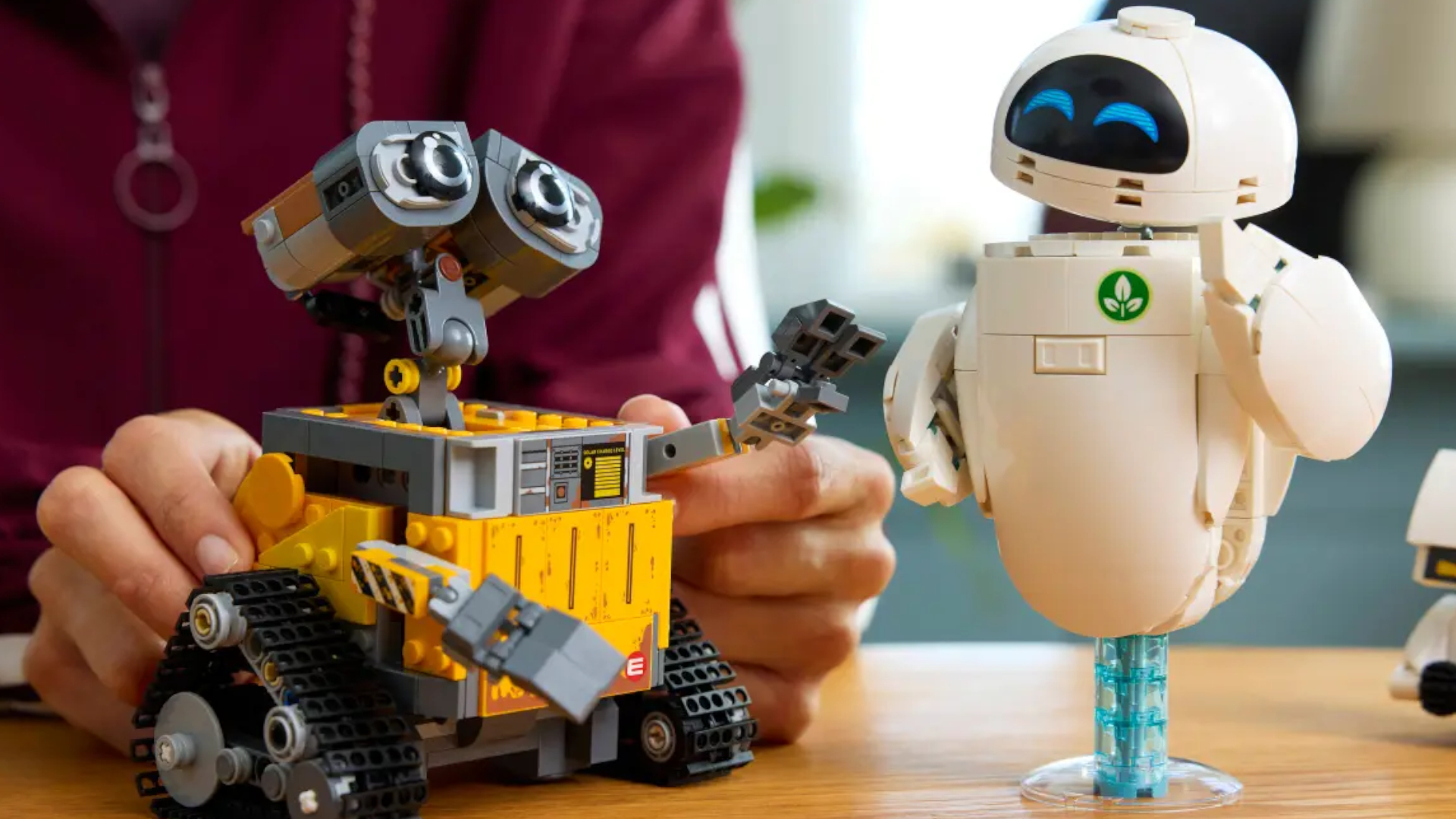 A hand moves Lego WALL-E&#039;s arm as the model sits on a wooden surface beside the EVE model